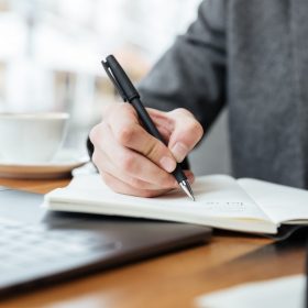 Cropped image of business man sitting by the table in cafe with laptop computer and writing something